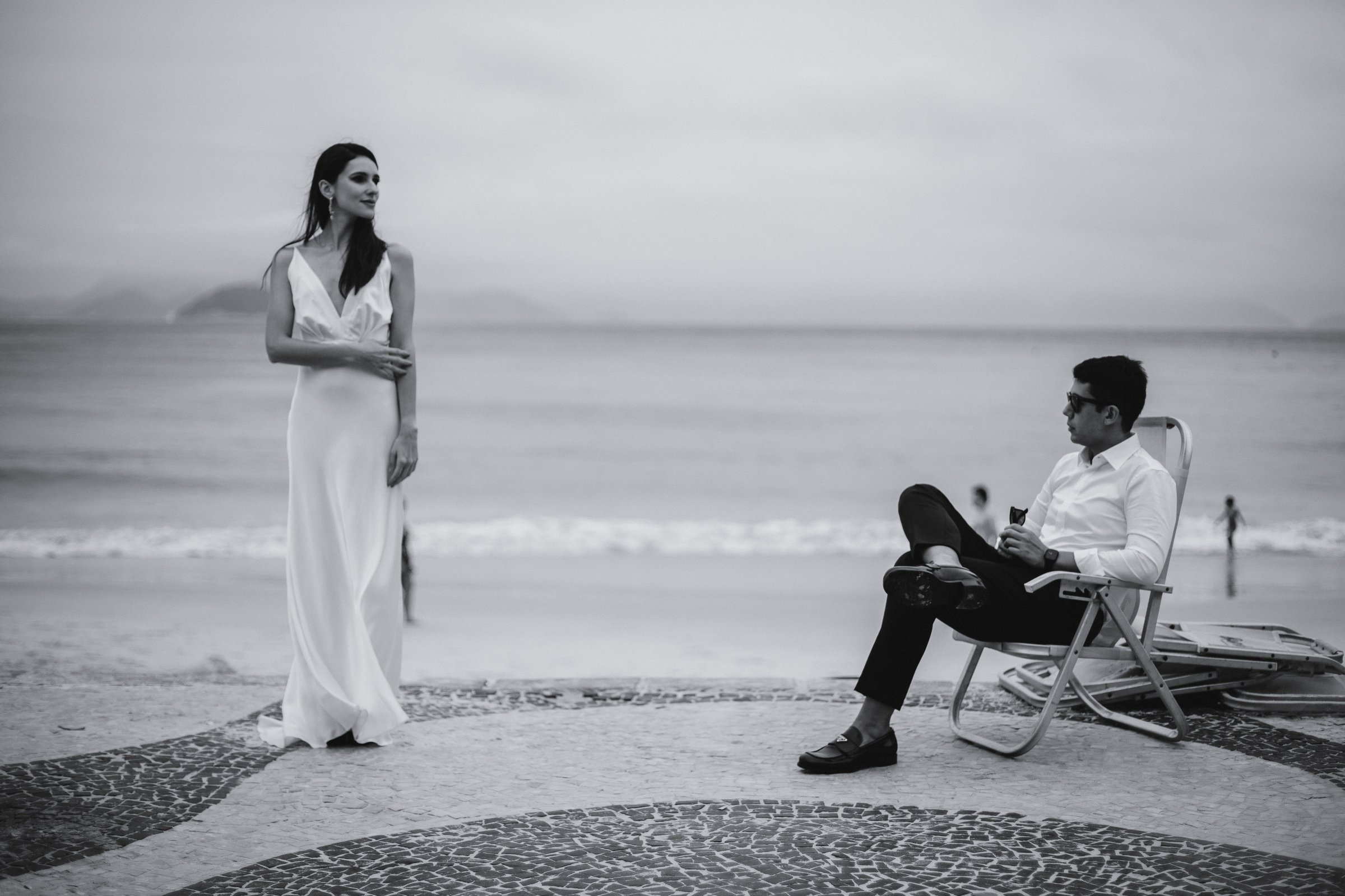 Casal em preto e branco, sentado à beira-mar, se olhando.