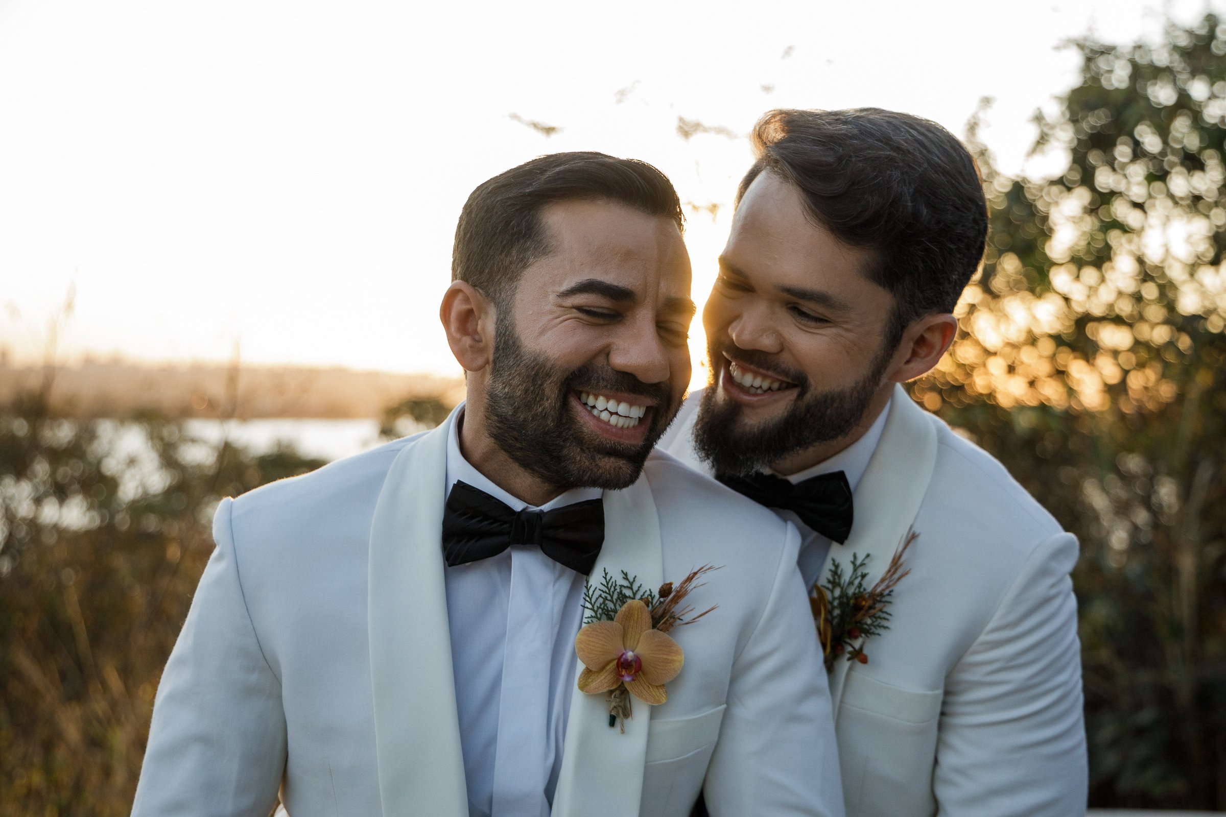 Dois noivos sorrindo um para o outro ao entardecer, vestindo terno branco com flor na lapela.