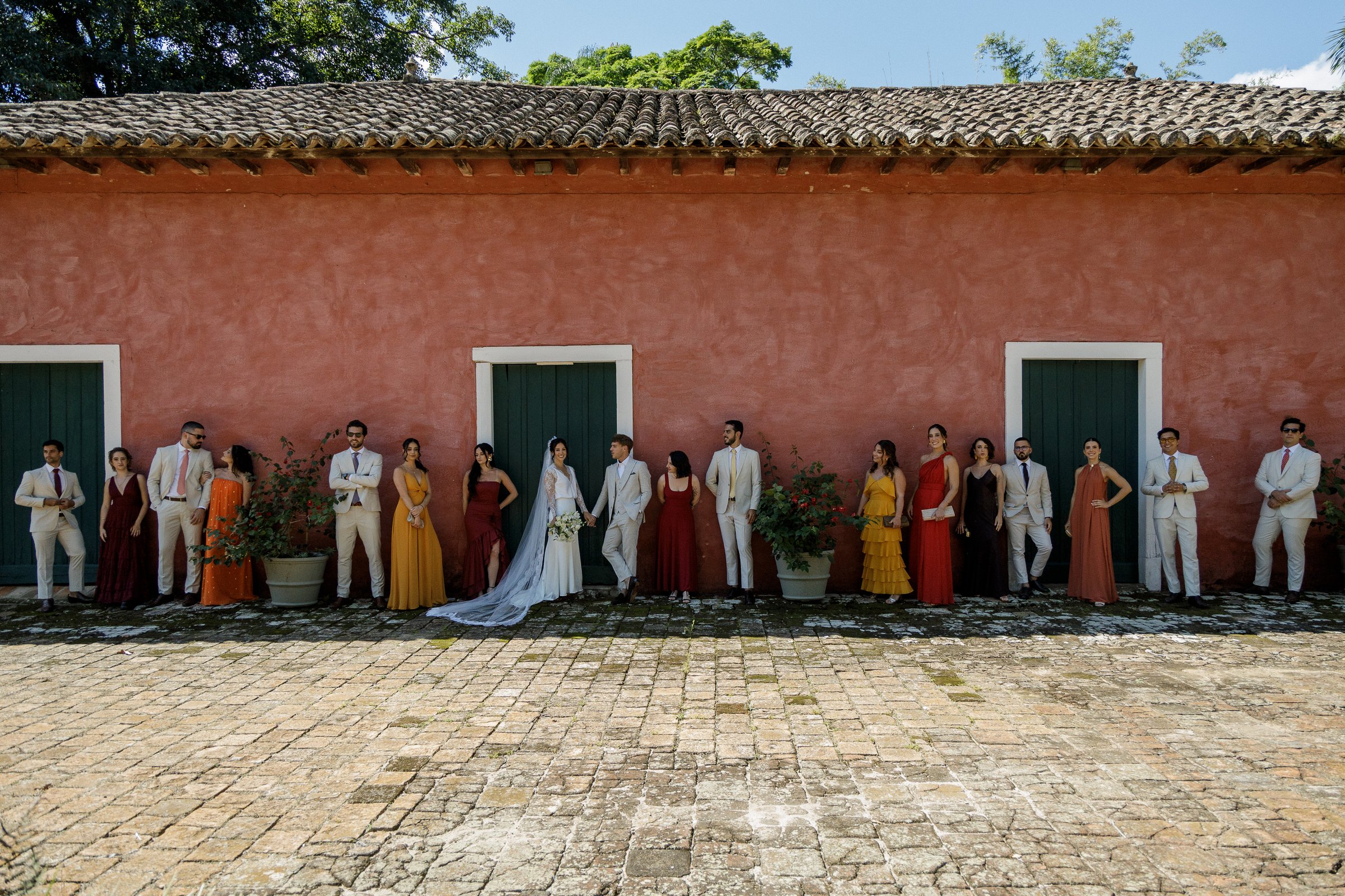 Padrinhos e noivos em fila na frente de um casarão rosa, durante a cerimônia.