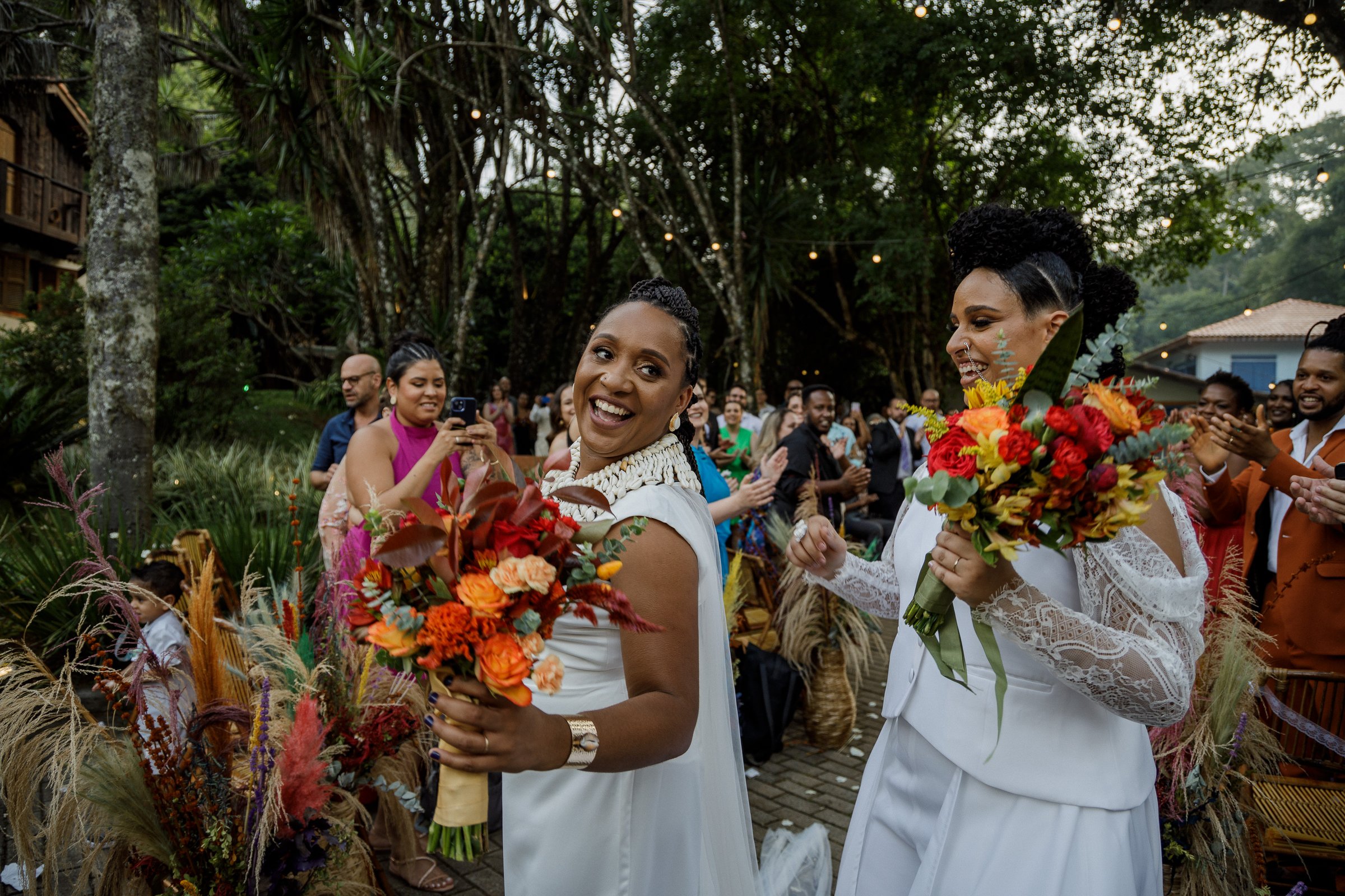Duas noivas sorridentes carregando buquês coloridos, cercadas por convidados numa procissão festiva.